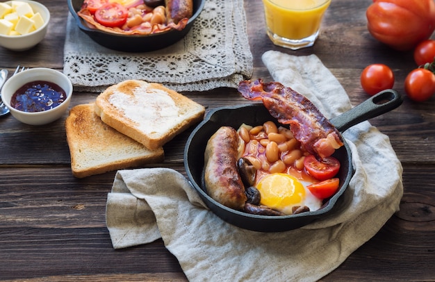 Fried eggs, sausages, bacon, beans and mushrooms in iron skillet, toasts, orange juice, butter and jam on rustic wooden background. Full english breakfast.