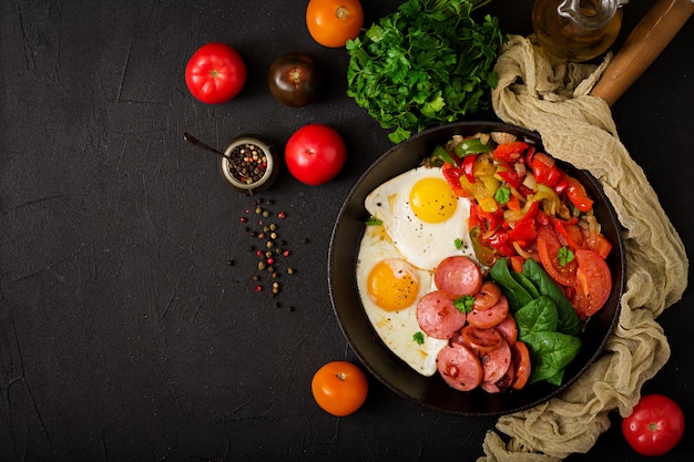 Fried eggs with sausage and vegetables in a frying pan on a black table in rustic style