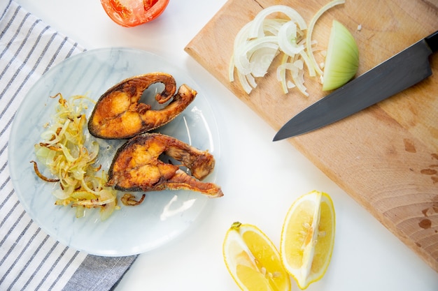 Fried fish with onions is on a plate on a waffle towel next to it is a cutting board with chopped on...