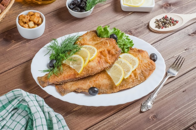 Fried flounder fish with lemon and herbs on a white plate on a dark wooden background.