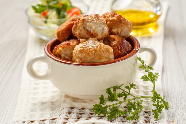 Fried meat cutlets in ceramic soup bowl, bottle with sunflower oil and salad of fresh cucumbers and tomatoes in glass bowl on white kitchen towel.