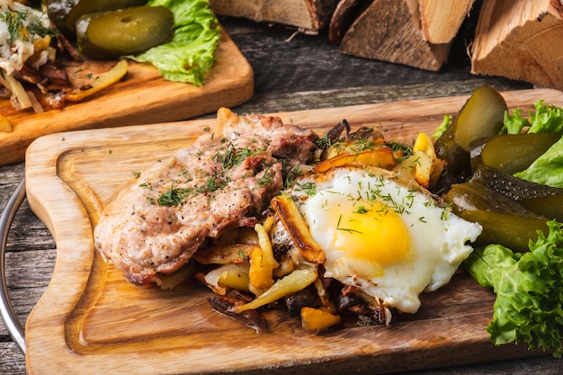 Fried meat with fried potatoes, eggs and salted cucumbers on wooden board.