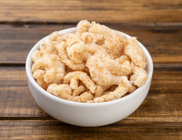 Fried pork cracklings on a white bowl over wooden table.