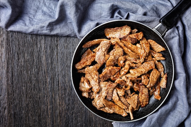 Fried ribeye beef steak cut into strips on a skillet on a dark wooden table, horizontal view from above, flat lay, free space