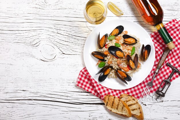 Fried rice with seafood mussels, shrimps and basil in a plate with wine bottle, wineglass, towel and toasted baguette on white cracked wooden table. Top view.