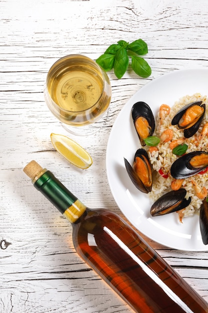 Fried rice with seafood mussels, shrimps and basil in a plate with wine bottle and wineglass on white cracked wooden table. Top view.