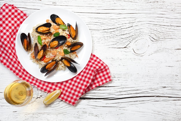 Fried rice with seafood mussels, shrimps and basil in a plate with wineglass, towel and toasted baguette on white cracked wooden table. Top view.