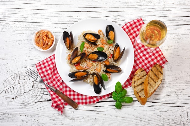 Fried rice with seafood mussels, shrimps and basil in a plate with wineglass, towel and toasted baguette on white cracked wooden table. Top view.