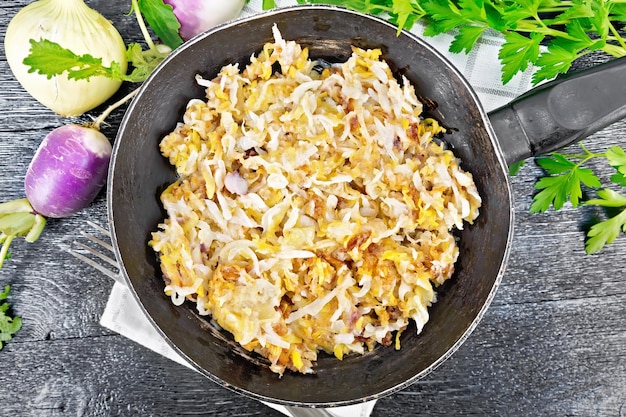 Fried turnips with onions in a frying pan on a towel on wooden board background from above