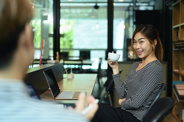 Friendly asian female employee talking with her colleague during coffee break in corporate office