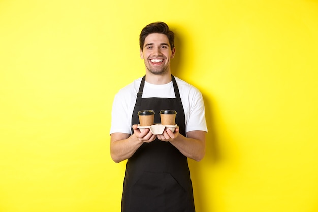 Friendly barista in black apron giving takeaway order, holding two cups of coffee and smiling, standing over yellow background.