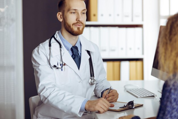 Friendly bearded doctor and patient woman discussing current health examination while sitting in sunny clinic. Medicine concept.