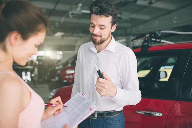 Friendly car salesman talking to a young woman and showing a new car inside showroom Signing of contract.