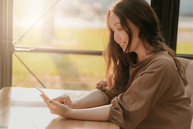 Friendly charming brunette woman with long curly hair sitting at the window in cafe with mobile phone in hands