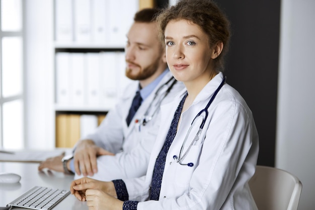 Friendly female doctor sitting in clinic with her male colleague at the background. Perfect medical service and medicine concept.