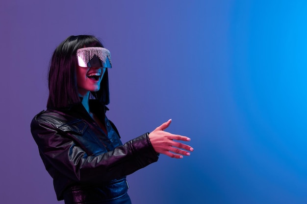 Friendly pretty awesome brunet woman in leather jacket trendy specular sunglasses greet someone posing isolated in blue violet color light background Neon party Cyberpunk concept Copy space