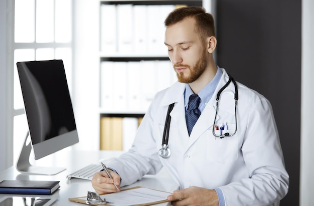 Friendly red-bearded doctor sitting and writing at clipboard in clinic. Perfect medical service in hospital. Medicine concept.