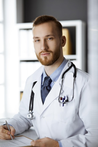 Friendly red-bearded doctor sitting and writing at clipboard in clinic. Perfect medical service in hospital. Medicine concept.