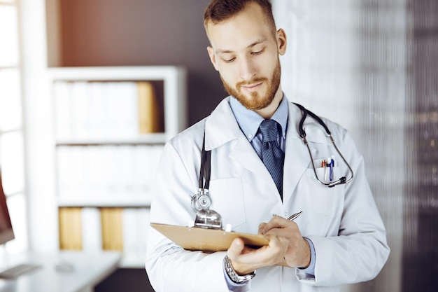 Friendly red-bearded doctor standing and writing with clipboard in sunny clinic at his working place. Medicine concept.