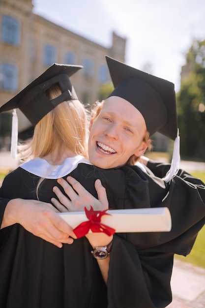 Friendly relations. Happy boy smiling in hugs of his groupmate standing in the university yard after their graduation.