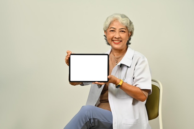 Friendly senior woman doctor in white coat holding digital tablet sitting against brown background