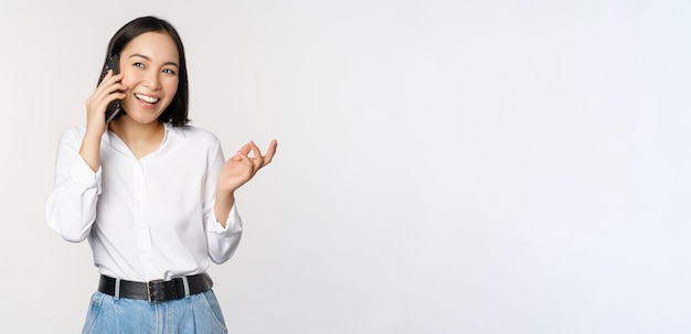 Friendly smiling asian woman talking on phone girl on call holding smartphone and laughing speaking standing over white background