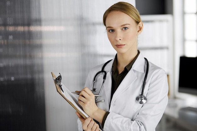 Friendly smiling female doctor using clipboard in clinic. Portrait of friendly physician woman at work. Medical service in hospital. Medicine concept.