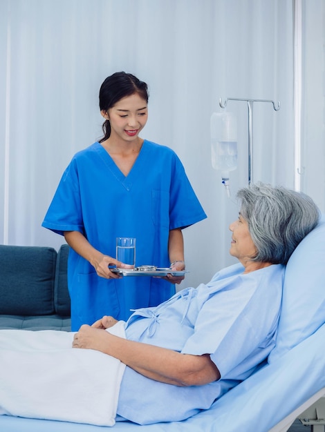 Friendly smiling young Asian nurse in blue scrub holding tray of pills for elderly patient lying on the bed in hospital room taking medicine or vitamin supplements senior healthcare and medical