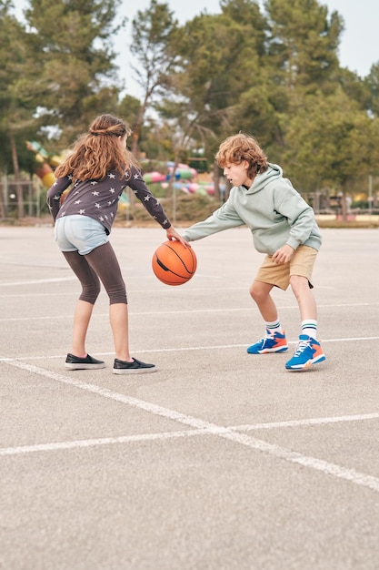 Friendly teenage girl and boy playing basketball on asphalt playground while entertaining together in summer