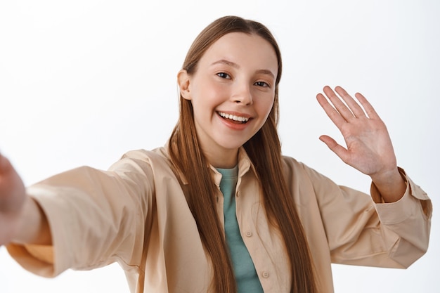 Friendly teenage girl waving at smartphone camera, video chat during covid-19 quarantine, stay in touch via mobile phone app, say hello, greeting you, standing over white wall