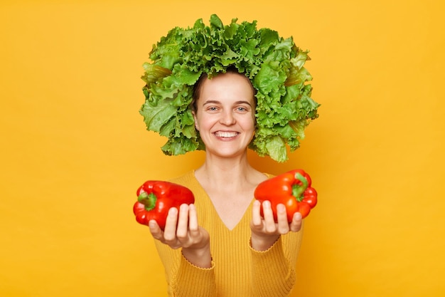 Friendly woman wearing casual shirt and lettuce wreath holding bell pepper standing isolated over yellow background offering fresh bell peppers looking at camera