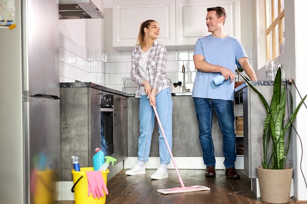 Friendly young couple cleaning home in kitchen woman wiping floor with mop husband helping