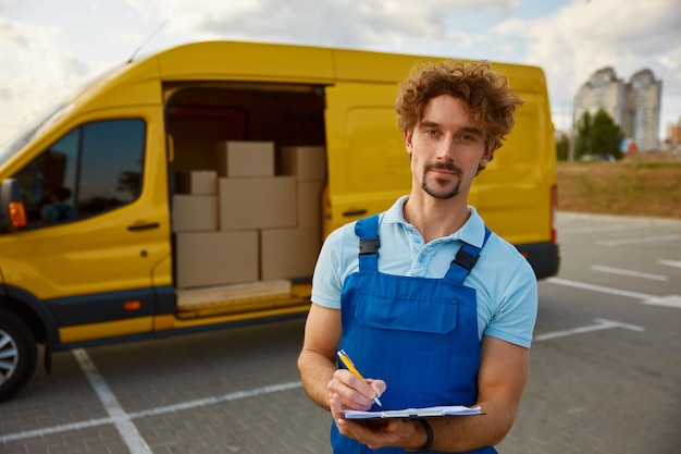 Friendly young courier with checklist and pen on clipboard Deliveryman standing nearby minivan full of cardboard packages Logistics worker portrait