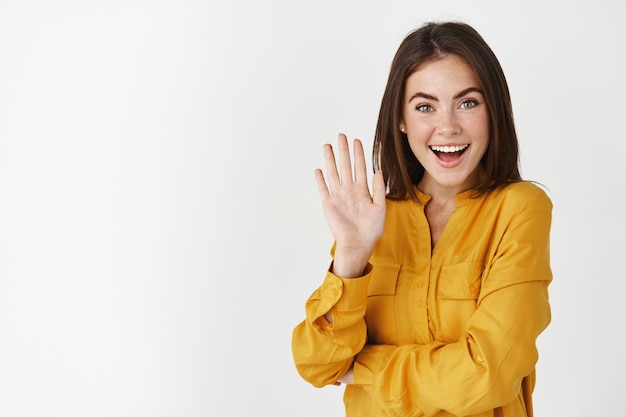 Friendly young woman waving hand and saying hi, greeting you with joyful smile, standing over white wall