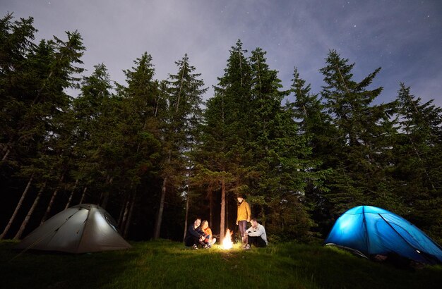 Friends around a campfire between tents amid powerful firs under a starry sky Nightcaping in the forest