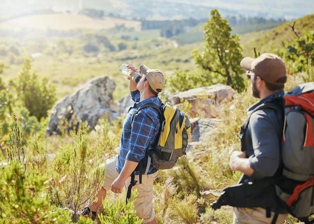 Friends hiking on grass in nature for fitness wellness and health as thirsty man drinking water and trekking in summer Men walking exercise and travel on rocky mountains for an adventure challenge