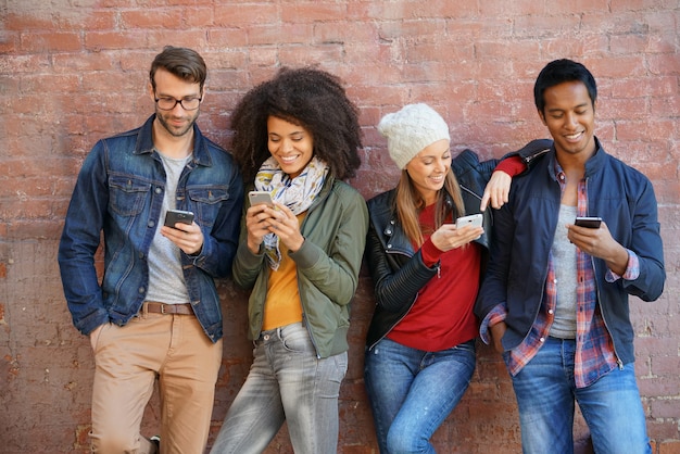 Friends leaning on brick wall, playing with smartphones