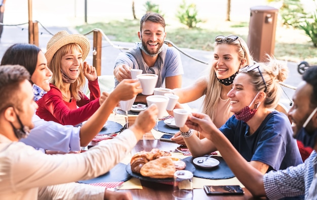 Photo friends toasting cappuccino at coffee shop wear face mask down - young people having fun together at restaurant - new normal lifestyle concept with happy guys and girls at cafe bar