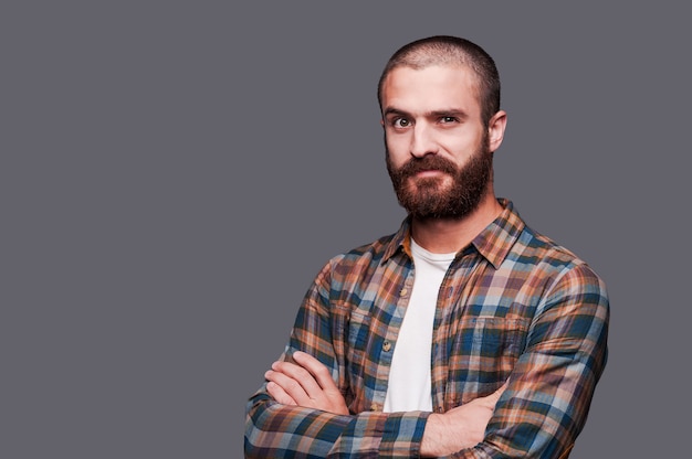 In friends with irony. Handsome young bearded man keeping arms crossed and expressing ironic smile while standing against grey background