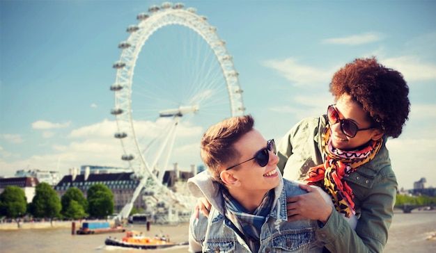 friendship, leisure, international, freedom and people concept - happy teenage couple in shades having fun over london ferry wheel background