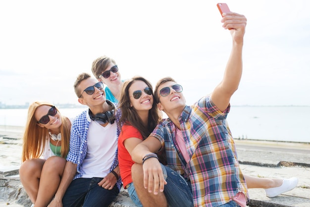 friendship, leisure, summer, technology and people concept - group of happy friends with smartphone taking selfie outdoors