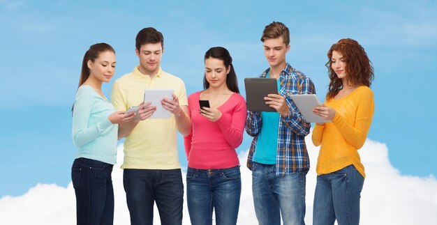 friendship, technolpgy and people concept - group of smiling teenagers with smartphones and tablet pc computers over blue sky with white cloud background