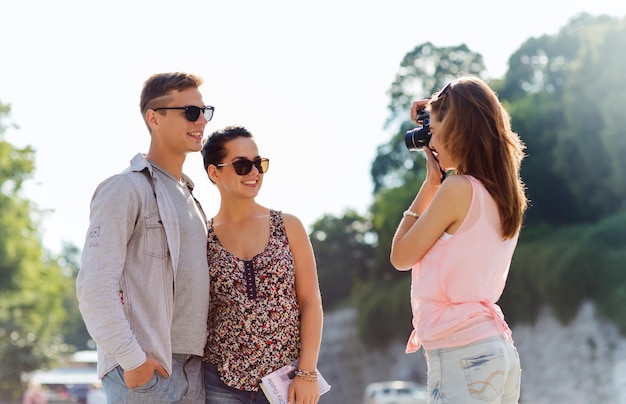 friendship, tourism, summer, technology and people concept - happy friends with camera taking picture outdoors