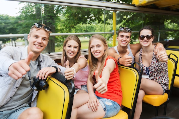 friendship, travel, vacation, summer and people concept - group of happy friends with digital camera traveling by tour bus and showing thumbs up