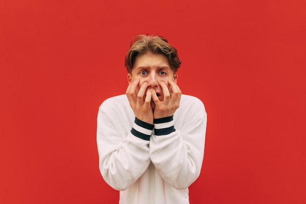 Photo frightened nervous guy in white clothes is isolated against a red wall covers his face