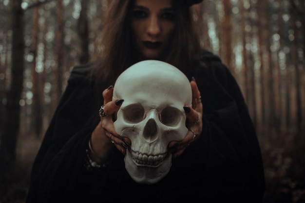 Frightening evil witch in black rags holds a dead man's skull in her hands for a dark ritual in the forest