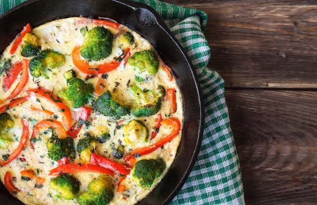 Frittata with broccoli and red pepper in iron skillet on rustic wooden table. Top view.