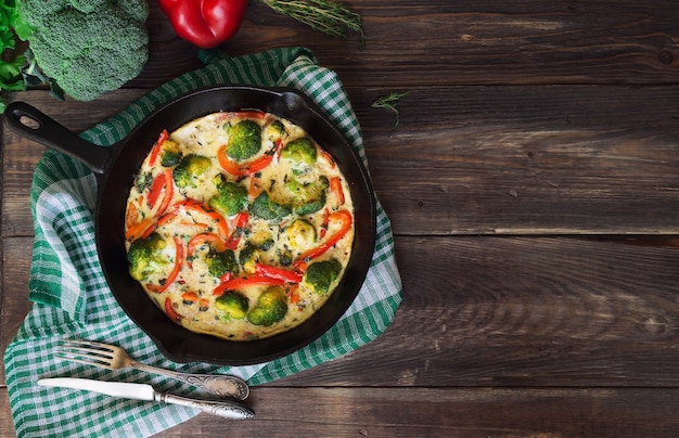 Frittata with broccoli and red pepper in iron skillet on rustic wooden table with ingredients. Top view. Copy space area.