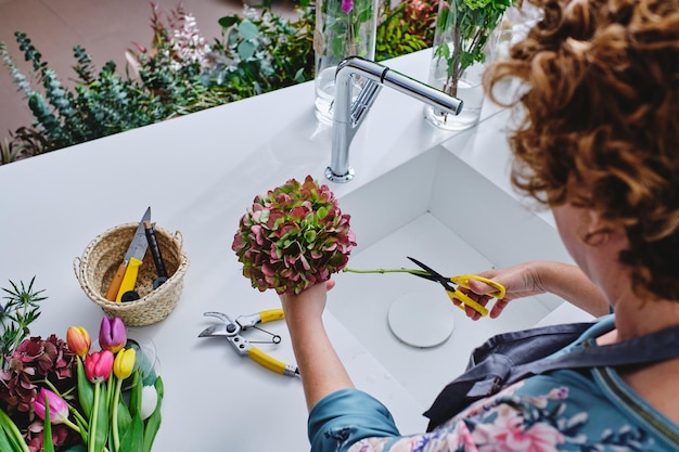 From above of anonymous female florist arranging bouquet of hydrangeas and cutting stem with scissors near sink in floral store