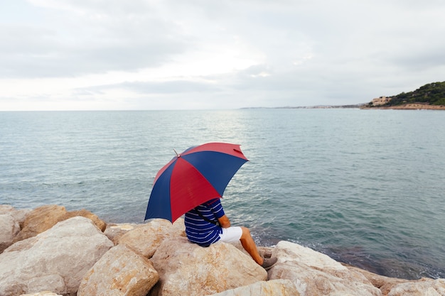 From behind anonymous man sitting on the rocks with an umbrella by the sea while it rains